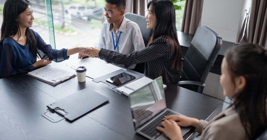 Business meeting with two people shaking hands across a table while others work on laptops.
