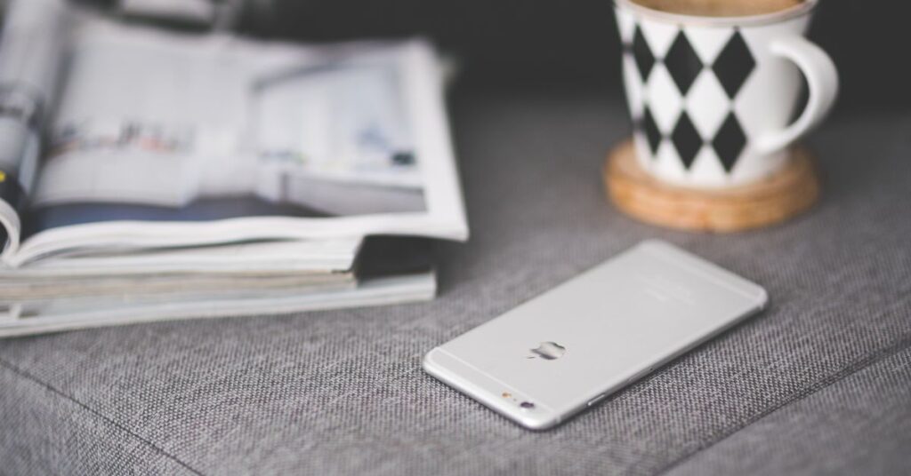 Smartphone placed on a gray fabric surface beside a coffee mug and an open magazine.