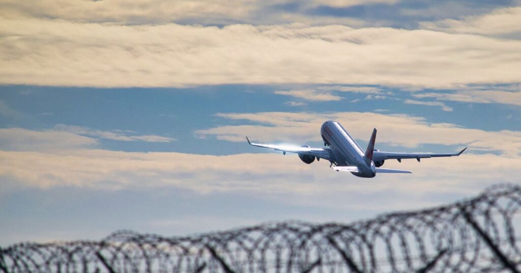Airplane taking off above airport fence and cloudy sky.