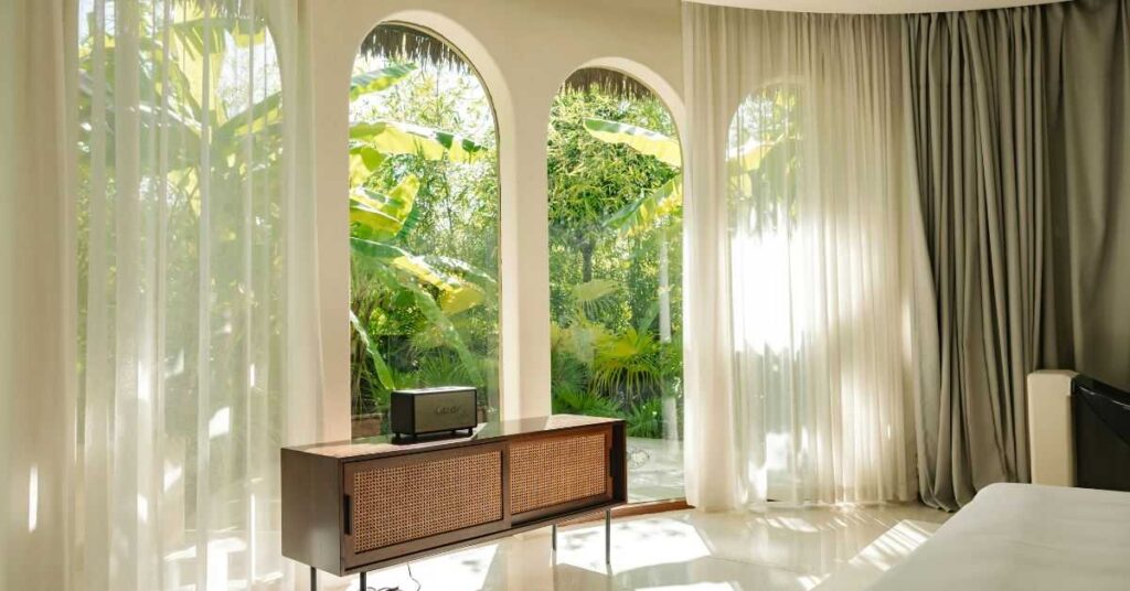 Bright and elegant living room with large arched windows, sheer curtains, and natural greenery visible outside.